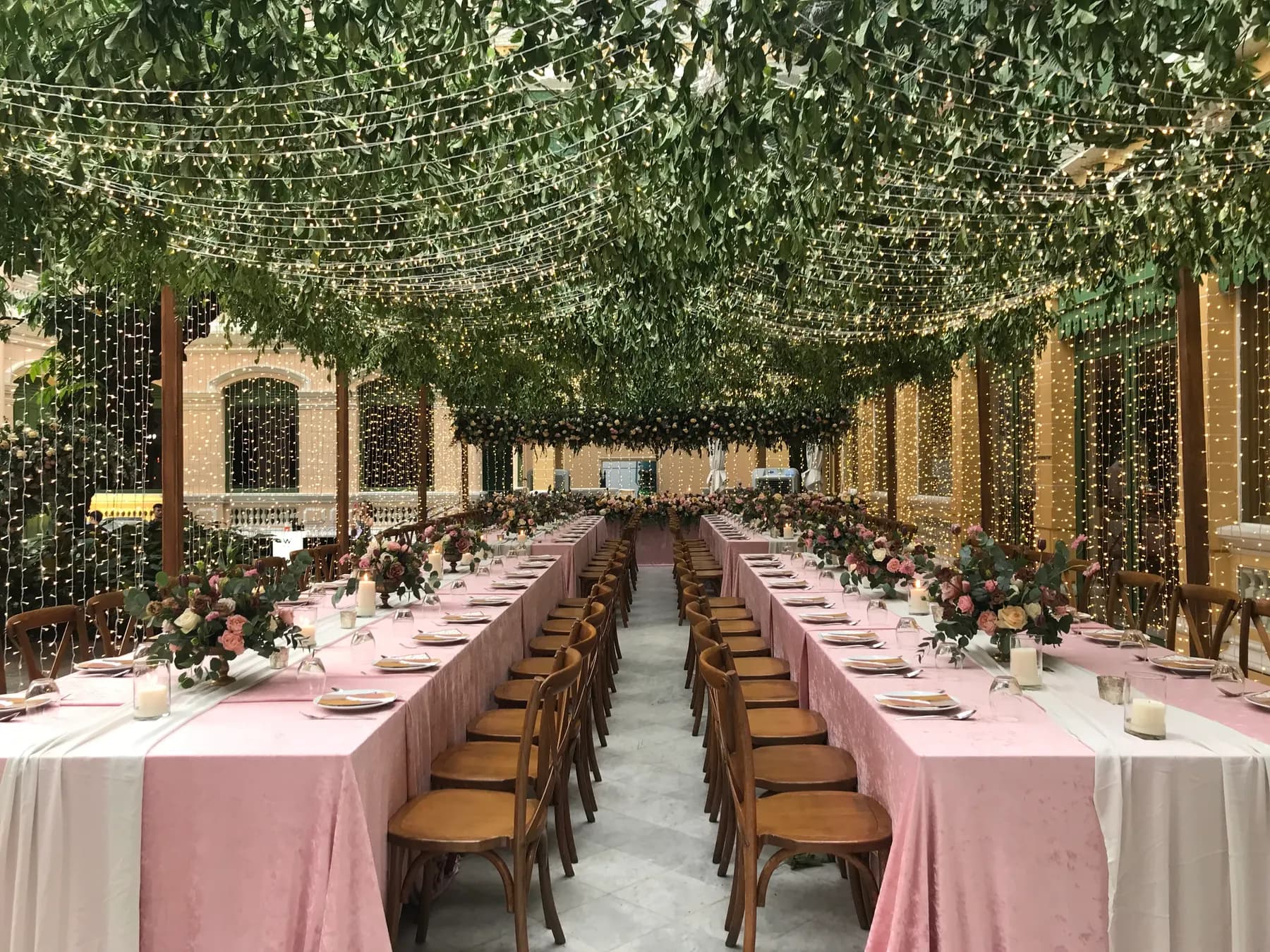 Long dinner tables with blush runners set beneath a canopy of greenery and fairy lights